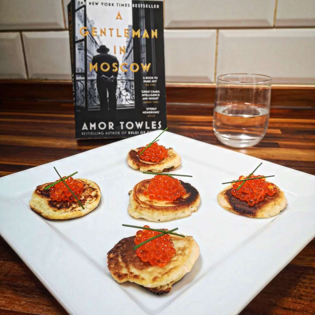 The book A Gentleman in Moscow next to very delicious looking blinis and a healthy sized pour of vodka